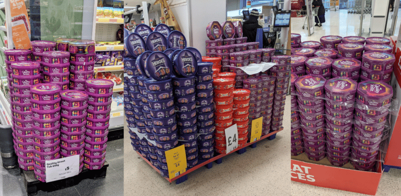 Stacks of seasonal purple tins of Heros, Celebrations, and Quality Street as a Christmas themed display in a U.K. grocery store
